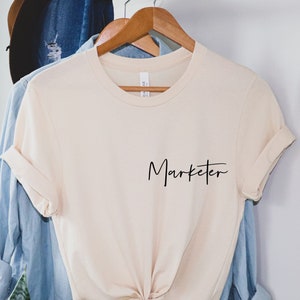 May include A light beige t shirt with the word Ma